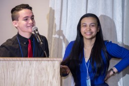 two students stand at a podium, one speaking into a microphone, the other smiling at the crowd