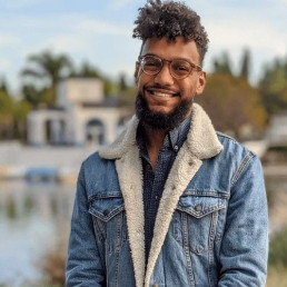 Headshot of Nicholas St. Fleur, wearing a blue denim jacket and smiling with a waterfront and palm trees in the background