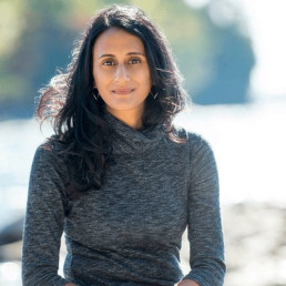 Headshot of Bina Venkataraman, smiling at the camera and wearing a gray dress, with a brightly lit waterfront in the background