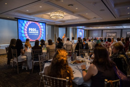 Guests seated at round tables face a projector screen that reads 