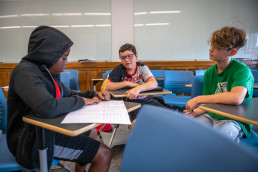 Three students sit at desks in a circle, poring over a piece of paper.