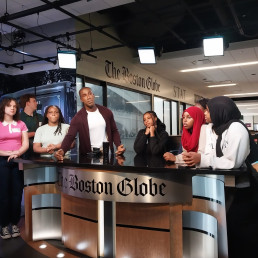 Five students and one reporter cluster around the Boston Globe TV desk