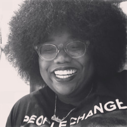 Black and white photo of a smiling woman with an Afro hairstyle and glasses is wearing a T-shirt that reads 